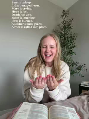 A woman with blonde hair laughs enthusiastically, facing the camera. She wears a white sweater over a light-colored top. Her hands are up, palms toward the viewer, as if offering something. An open book, likely a bible, with visible text, rests on the surface below. The background includes a blurred room setting with an indoor plant.