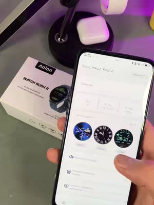A person holds a smartphone displaying a smartwatch app interface, featuring various watch faces. To the left, a smartwatch box sits on a table, alongside a charging stand with a pair of white earbuds. The scene is well-lit with a focus on the technological setup.