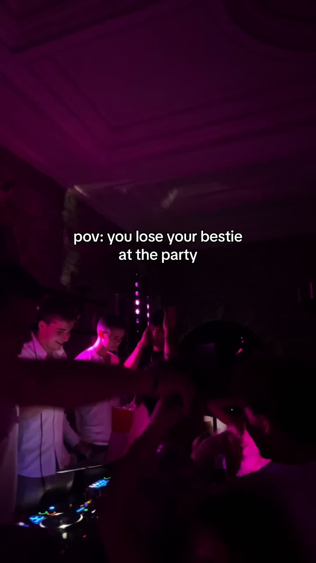 The image shows a dimly lit party scene. In the foreground, partially visible figures, likely attendees, suggest a crowded environment. Soft pink lighting bathes the space. In the background, more partygoers are visible as well. The text overlay reads, "pov: you lose your bestie at the party".