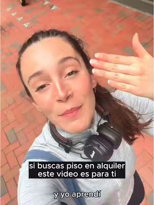 A woman is looking directly at the camera, with her right hand held up near her face. She is wearing headphones and the background is a brick-patterned ground.
