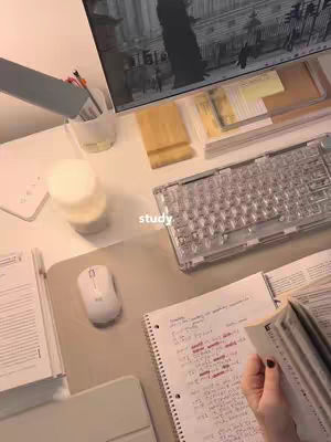 An overhead shot of a well-organized desk set up for studying. A large computer monitor displays a street scene in grayscale, and a clear mechanical keyboard and a white mouse are positioned on the desk. Several notebooks and open books are scattered around, and a hand is visible holding a book open, hinting at active study. The color palette is muted, with a focus on shades of white and beige, creating a calm and organized atmosphere.