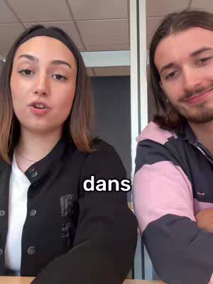A split-screen image featuring a young woman on the left and a young man on the right. The woman is positioned closer to the camera, facing the viewer and speaking. Her hair is shoulder-length and dark, and she wears a black headband and a black jacket. A white text overlay appears on the image: 'dans' (in French). The man is positioned further back from the camera, with brown hair, a shorter beard, and he is smiling, appearing attentive. The background is out of focus, suggesting an office environment.