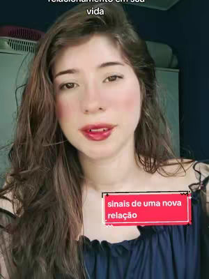 A woman's face is center in the frame, with long brown wavy hair. She has a slightly open mouth with red lipstick. A red rectangular overlay with white text is in the lower right corner. The background is a dimly lit room.