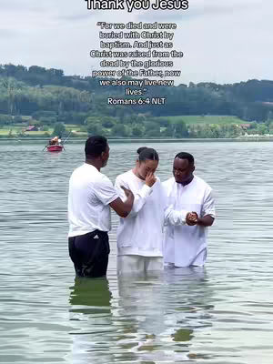 Three people standing in water during a baptism ceremony. The woman in the middle has her eyes closed. The background shows a serene lake and lush green hills.
