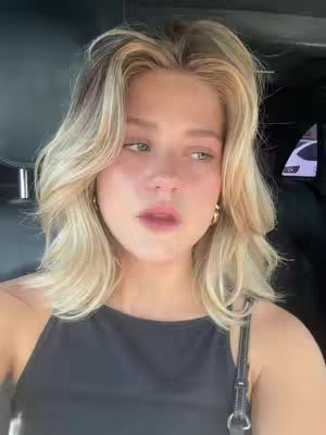 A young woman with wavy blonde hair is seen from the chest up, framed in what appears to be a car interior. She wears a black tank top and gold hoop earrings. Her expression is thoughtful, her gaze slightly to the side, with hints of subtle makeup around her eyes and lips. The natural light suggests a daytime setting.