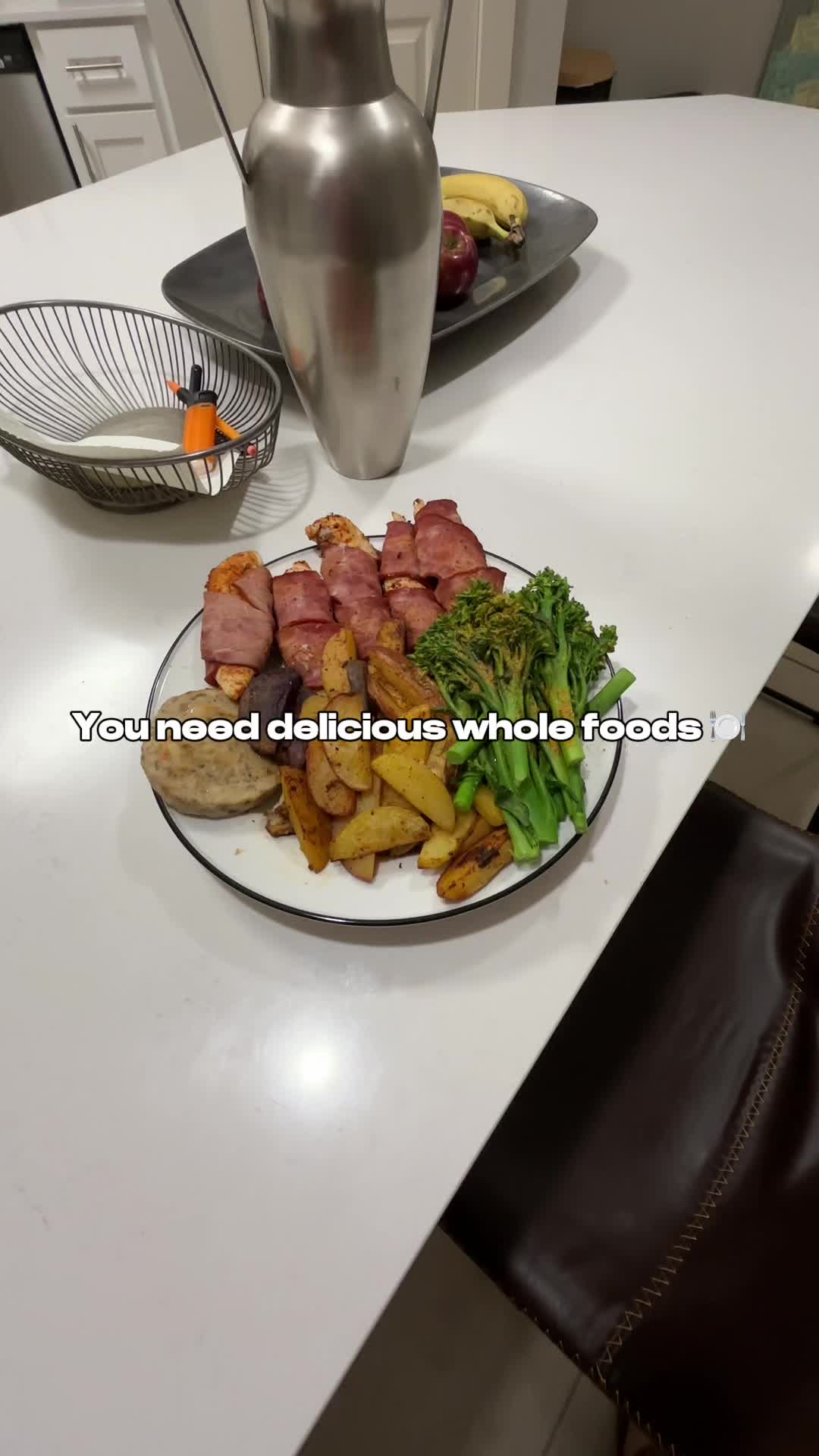 A photo showcasing a plate of food set on a white surface. The plate holds several food items, including what appears to be chicken wrapped in bacon, alongside roasted potatoes, broccolini, and a single, large, cooked potato. The background includes elements indicative of a kitchen or dining area, such as a countertop, a metal pitcher, a wire basket, and a plate of fruit.