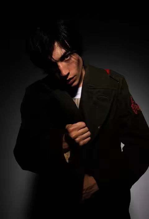 The cover image features a young man in the foreground, shot from the chest up, against a dark gray backdrop that fades to black. He is wearing a dark green military-style jacket with a prominent red marking on the sleeve. His hair is dark and tousled, and his gaze is directed at the camera with an intense expression. The lighting is dramatic, highlighting his face and upper body, and the overall composition is minimalistic and focuses on the figure.