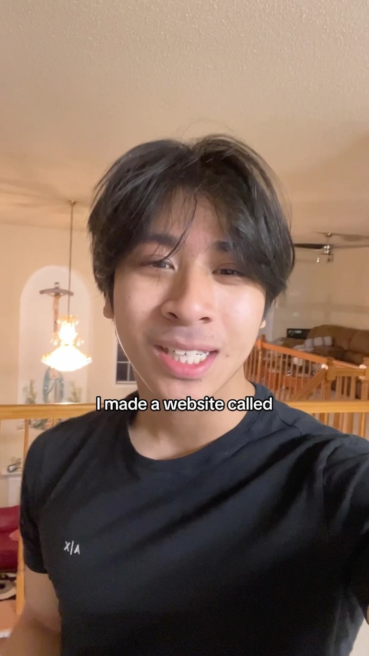 A young man is centered in the frame, smiling at the camera with a warm expression. He has dark hair styled away from his face. He wears a black t-shirt. Behind him, a casual indoor setting is visible, with a chandelier and other architectural details.