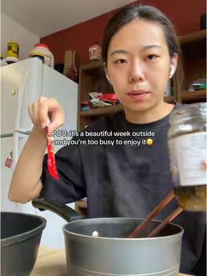 A woman in a kitchen setting. She is holding a red chili pepper and is looking at the camera with a slightly upset expression. The background includes a refrigerator, shelves, and a pot on the stove.