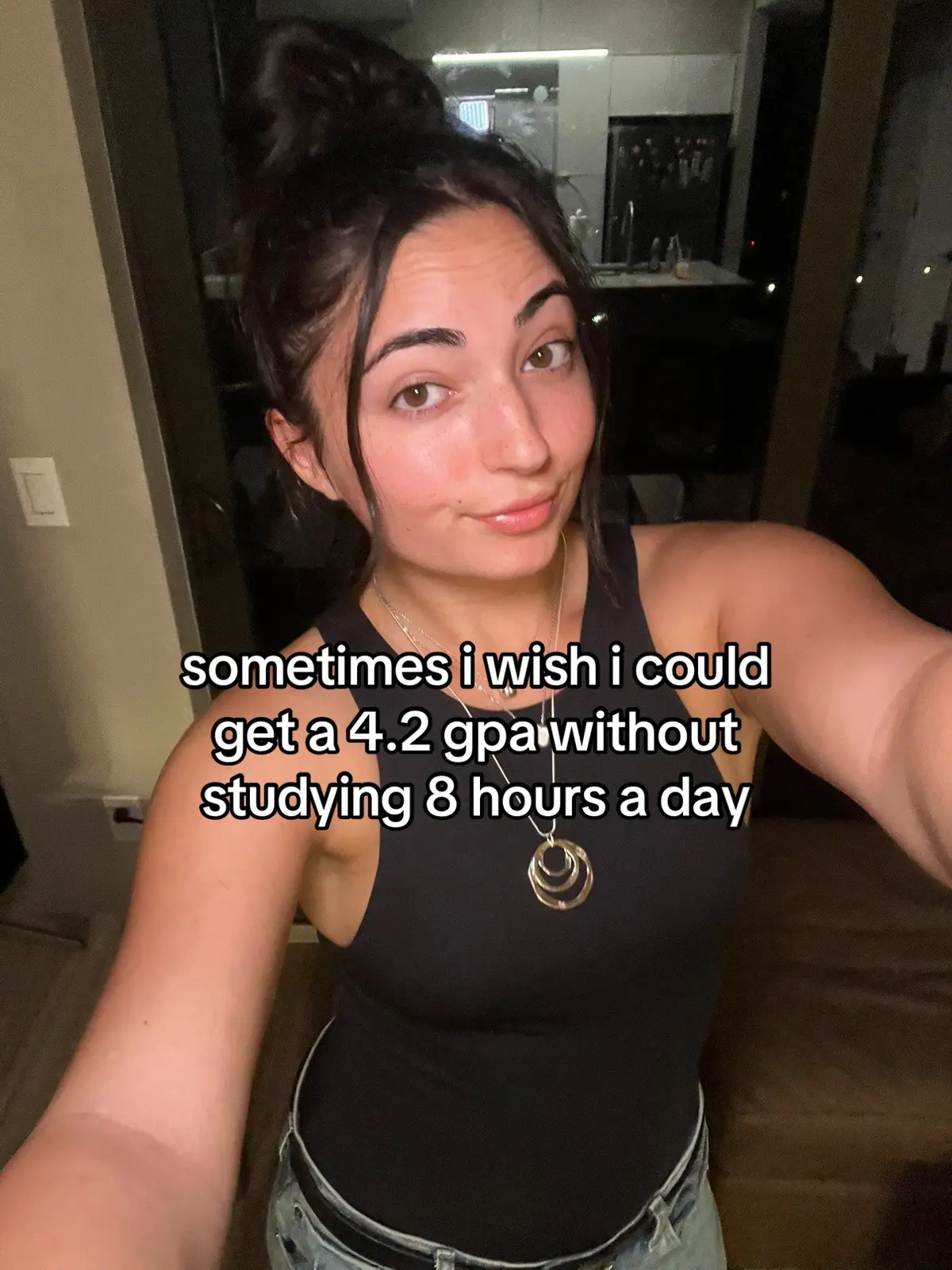 A selfie-style shot featuring a young woman against a softly lit, dark backdrop. She is wearing a black tank top and has her dark hair pulled up in a bun. Overlaid text in white font states, 'sometimes i wish i could get a 4.2 gpa without studying 8 hours a day'. The woman is looking at the camera, with a slight smile and a thoughtful expression. The lighting seems to be coming from a source above and left of the frame, casting gentle shadows. The composition is typical of a selfie, drawing direct attention to the subject.