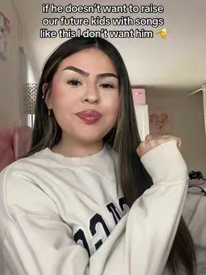 A young woman with long dark hair and makeup is centered in the frame, slightly smiling while looking towards the bottom right corner. The top of her sweater, which bears a large letter, is visible. The background is blurred, suggesting an indoor setting, possibly a bedroom with a decorated wall and a glimpse of other furniture.