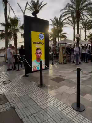 A poster of a man wearing sunglasses is propped up in front of an outdoor setting. There are people and palm trees in the background.