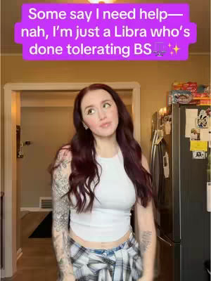 A young woman with long auburn hair is standing inside a home. She's wearing a white tank top, a plaid shirt tied around her waist, and jeans. The background includes a doorway, a refrigerator, and a glimpse of what appears to be outside. A text overlay at the top of the image reads, \"Some say I need help - nah, I'm just a Libra who's done tolerating BS.\"