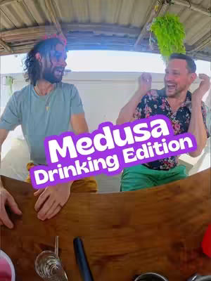 The cover image shows two men sitting at a wooden bar or table, both smiling and looking at each other. The man on the left has long hair, sunglasses, and a blue shirt. The other man on the right has short hair, a floral shirt, and green shorts. A purple text overlay reads "Medusa Drinking Edition". Behind them is a blurred-out, natural-looking setting.