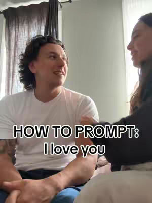 A man and a woman are facing each other, seemingly in a conversation, with the man in the foreground wearing a white t-shirt, and the woman partially visible on the right side. The background is a room with a curtain and window.
