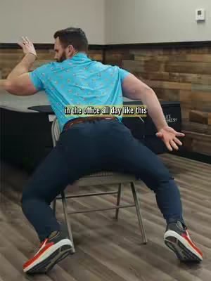 A man is leaning back in a chair, with his legs spread wide. He's turned away from the camera, towards the right. The background includes a simple, light-colored wall with a few details and what looks like a window or a large item of furniture behind him.