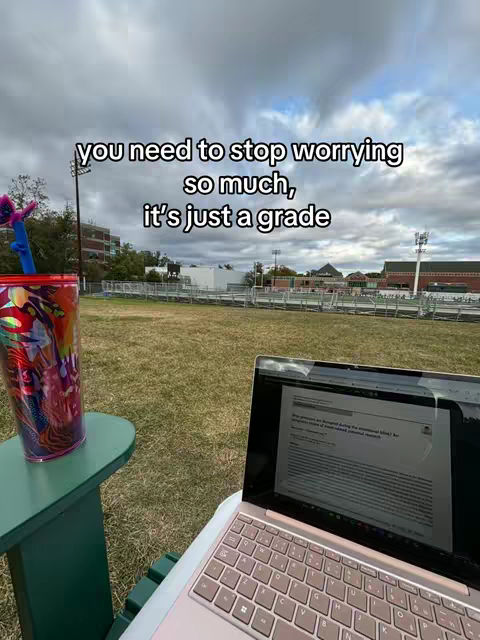A laptop is open on a chair with a green tabletop next to a colorful cup. The background is a cloudy day with grass and some distant buildings.
