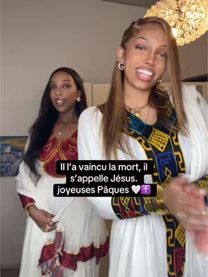 Two women are posing for a picture, likely in a home setting, close to the camera. The woman on the right is in the foreground, smiling and looking directly at the camera. She has light brown hair, and is wearing a white dress with a gold and intricate design around the upper part. The woman on her left, in the background, is wearing a similar dress with a decorative red pattern and has darker hair. Both women have a warm skin tone. The text overlay declares 'Jesus has risen' in French with a greeting for Easter. The background is softly lit, featuring hints of interior design.