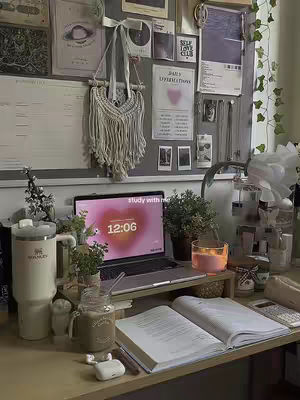 A well-lit, cozy desk setup with a laptop displaying a clock that reads 12:06 with a heart design. The desk also holds a Stanley tumbler, an open book, a mug, and other study-related supplies against a background of a bulletin board with notes and decorations, a candle, and plants.