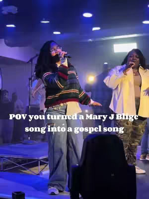 The image features a live music performance inside a venue. Two women stand in the foreground, each holding a microphone. The woman on the left wears a striped sweater, jeans, and glasses, and the other woman is to the right of her and dressed in a white jacket and camo pants. The stage is lit with blue lights, and the audience is partially visible in the background. Text overlayed on the image reads, 'POV you turned a Mary J Blige song into a gospel song.'