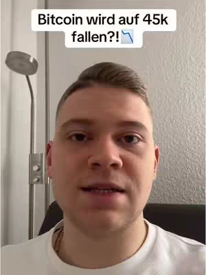 A close-up shot of a man looking directly at the camera. Above him, there is a white rectangular text overlay that reads 'Bitcoin wird auf 45k fallen?!', and a downward pointing arrow icon. The background consists of a plain wall, a small section of what looks like a metal lamp is visible on the left. The man is wearing a white t-shirt and a golden chain can be briefly seen.