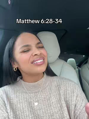 A woman is seated in a car, with the camera angled slightly upward, filling the frame with her face. She wears a tan sweater and a gold necklace with a heart pendant. Her dark hair is parted to the side and brushed away from her face, showing hoop earrings. Directly above her head is text that says "Matthew 6:28-34". The background is blurred, suggesting she is in motion, with elements of a car interior like the headrest and seat.