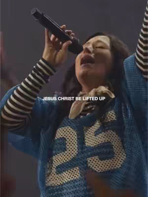 A woman with dark hair singing into a microphone with her arms raised. She wears a blue jersey with the number 25 and a black and white stripe sleeve. The background is dark but appears to be a stage.