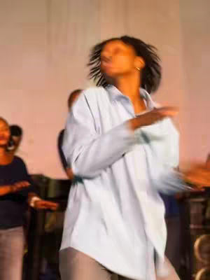 The image shows a blurred, dynamic scene featuring a person with dark hair in motion, seemingly dancing or performing. The individual, wearing a light blue button-down shirt and gray pants, is the focal point, with their arms outstretched. The background is indistinct, possibly capturing an indoor setting, like a stage or event where motion is emphasized. The overall effect is one of energy and movement.