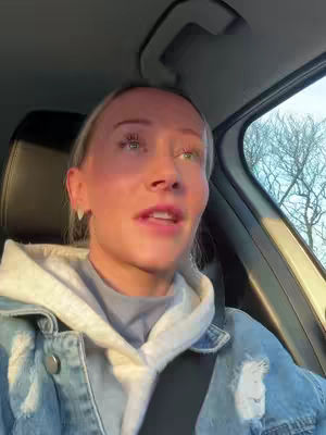A selfie taken from inside a car, showing a young woman with blonde hair and a denim jacket. The woman is looking up and slightly to the right, and the background includes the car's interior and a view of leafless trees through the window. The lighting suggests it's near sunset. The composition is centered on the woman's face and upper body.