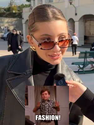 A woman wearing sunglasses and a leather jacket is holding what seems to be a microphone. A meme of a child with open arms with a black and white text overlay of "FASHION!" is overlaid.