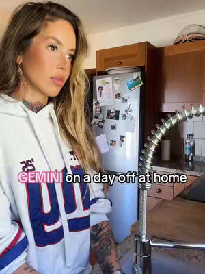 A woman in a white hoodie is in a kitchen. She is looking off to the side, the kitchen has wooden cabinets, a refrigerator with pictures on it, and a silver faucet.