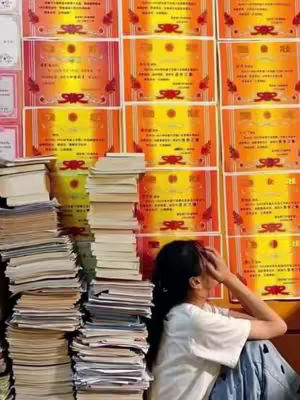 The image shows a young woman sitting on the floor with her head in her hand, appearing stressed or overwhelmed. She is surrounded by stacks of books and papers. Behind her is a wall covered in achievement certificates, suggesting a focus on academics and achievement. The color palette is dominated by yellows and oranges from the certificates and neutral tones elsewhere, creating a contrast that draws attention to the student and the environment.