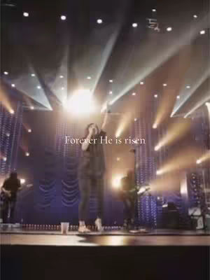 A live music performance on stage with a singer in the center under bright lights. Additional musicians are visible in the background on either side. Text overlayed on the image, with a warm color palette.