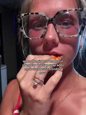 A selfie-style cover image features a fair-skinned woman with blonde hair and large, patterned-rimmed glasses. She is partially obscured by the frame, but her face and upper body are visible. She is holding a snack in her hand, with her fingers in her mouth. The expression is one of thoughtfulness. A ring with a large gem adorns her finger. Text overlayed in the lower part of the image reads: "when the girl who made fun of me for deleting snap adds me locket bc it's trending now".