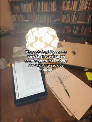 The image shows a study setup with a lamp with a cloud-like shade over a desk covered with a tablet, papers and a pen. The background includes bookshelves filled with books.