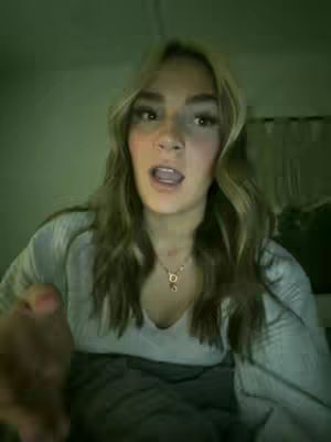 A woman with long, wavy blonde hair looks directly at the camera with an open mouth, as if speaking. She has visible eye makeup and is wearing a light-colored top and a necklace. The background is dimly lit, with a green hue suggesting the lighting in the room.