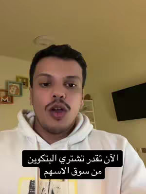 A selfie-style shot featuring a man in a white hoodie. He has a mustache and is speaking directly to the camera. A black rectangle at the bottom contains Arabic text. The background is a plain wall with some pictures and a television.