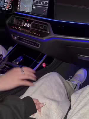 The image shows the interior of a car. The interior is dark with blue ambient lighting. In the foreground, the legs of two individuals are visible; one wearing gray sweatpants with a white shoe and the other with a black jacket and a silver ring. A hand with painted nails holds another hands in the foreground. The car's dashboard and infotainment system are visible, with a navigation screen in the background.