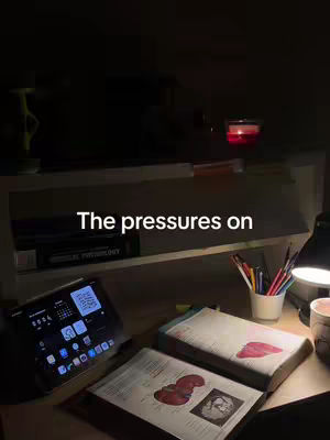 A dimly lit desk setup for studying, featuring an open textbook with anatomical illustrations, a tablet displaying a home screen, pens in a holder, and a lit candle. Text overlayed on the image reads, 'The pressures on'.