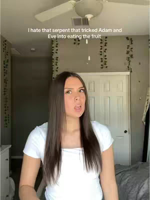 A woman with long brown hair is centered in the frame, looking slightly to the right and speaking with an open mouth. She wears a white t-shirt and stands against a backdrop of a bedroom with a ceiling fan, a door, and decorative vines.