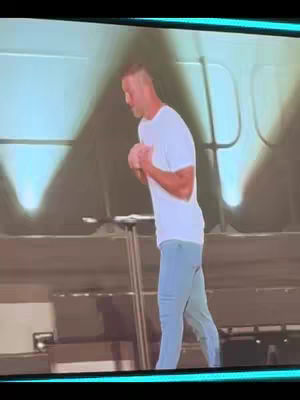 The image shows a man in a white t-shirt and light blue pants standing on a stage, holding his hands in front of him as if praying or speaking. The background is a blurred image of a large screen or wall, providing context of a public speaking event or presentation.