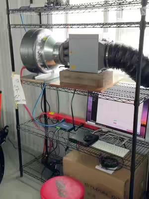 An indoor shot captures a mining rig setup on metal shelving. A large, cylindrical component with a vent is connected to a grey box, with black tubing. On the shelf below is a computer monitor displaying charts and graphs and a keyboard.