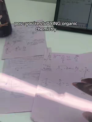The image shows a POV shot of someone studying organic chemistry. Piles of handwritten notes and diagrams fill the frame, with a hand pointing to a spot on the paper. A water bottle is on the left, while a laptop or tablet with a screen displaying text is in the top right corner. The lighting appears to be a mix of natural and artificial, creating shadows and highlights.