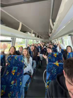 An interior shot of a bus filled with excited people. The focus is on the expressions of the individuals, who are waving their hands and smiling. The lighting suggests a bright, overcast day. The bus interior features patterned seats and overhead compartments, with the background blurred due to depth of field.