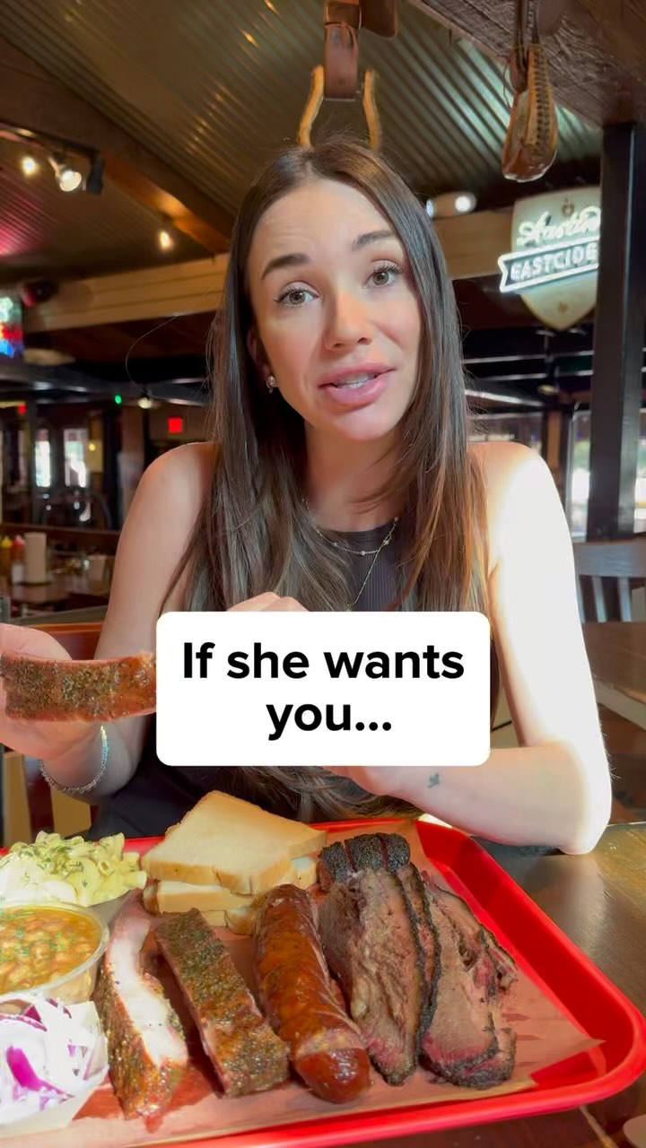 A woman with long brown hair is seated in a restaurant. She's holding a piece of brisket and wearing a dark top, speaking directly to the camera. In the foreground is a plate of food, including brisket, sausage, ribs, coleslaw, and beans, with the restaurant's decor visible in the background, including wooden furnishings and a sign.