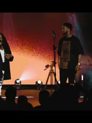 The image shows two people on a stage with microphones. The background is a mix of orange and red hues, possibly from stage lighting. There is a man on the right wearing a black t-shirt, and a woman on the left in a dark blazer, with silhouettes of people in the audience in the foreground.