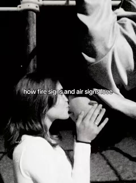 The image is a black and white shot of a person kissing another on the cheek. The person doing the kissing is on the left and appears to be a woman wearing a white shirt. The other person’s face is partially hidden. The background is dark and out of focus.