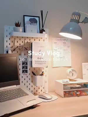 A well-lit desk setup with a laptop, notes, and decorative items. A lamp shines down on the desk, which also has a notepad and a small clock. Behind it is a white memo board with stationary and some images.