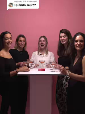 Five women are gathered, appearing in a vertical medium shot against a solid pink backdrop. They are positioned around a rectangular white table. On the table are glasses with pink liquid. The woman in the center is laughing and holding a glass. The others are smiling. One woman is wearing a white blouse and the others are wearing a variety of black tops. The background pink is the main color.