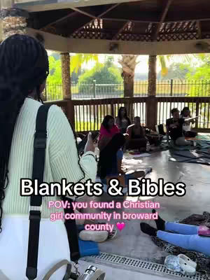 The image shows a woman from the back, in front of an outdoor gathering. The woman stands near the foreground, partially obscuring the view with her dark hair and a sweater, and a black strap over her shoulder. In the background, under a shaded structure, several people are seated on blankets, giving a sense of community and shared activity. The color palette is natural with green grass and sunlight.